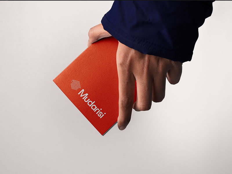 Mudarisi thumpnail A hand wearing a dark blue sleeve holds a red notebook titled