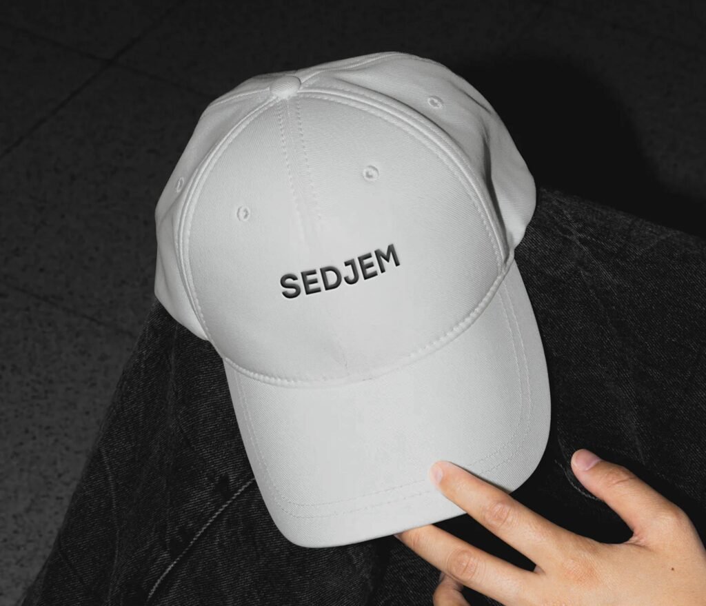 A white baseball cap with the word 'SEDJEM' embroidered in bold black letters on the front. The cap is held by a hand with visible fingers positioned at the bottom right corner of the image. The background features a dark, textured surface.